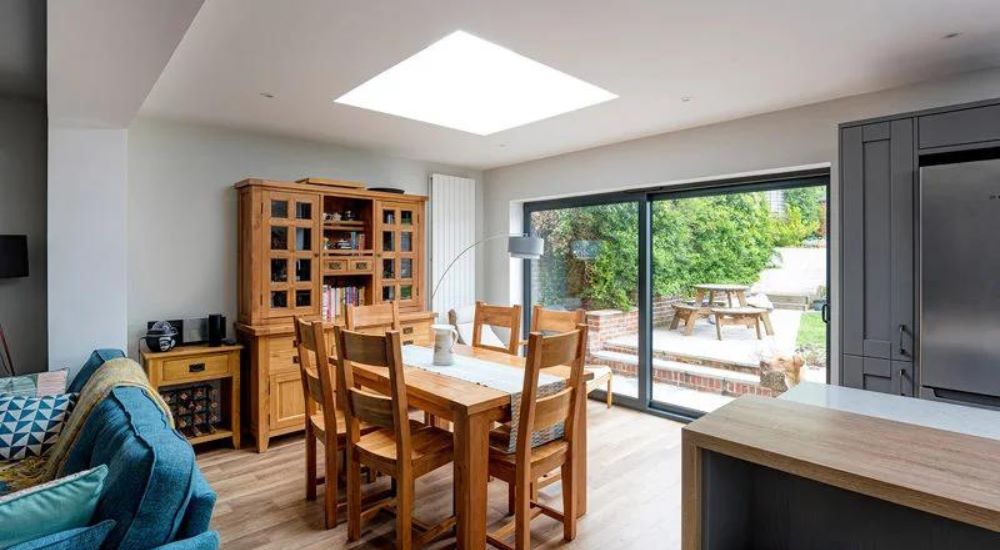 flat rooflight installed over wooden dining table and chairs, blue sofa to the left and patio doors to a garden to the right