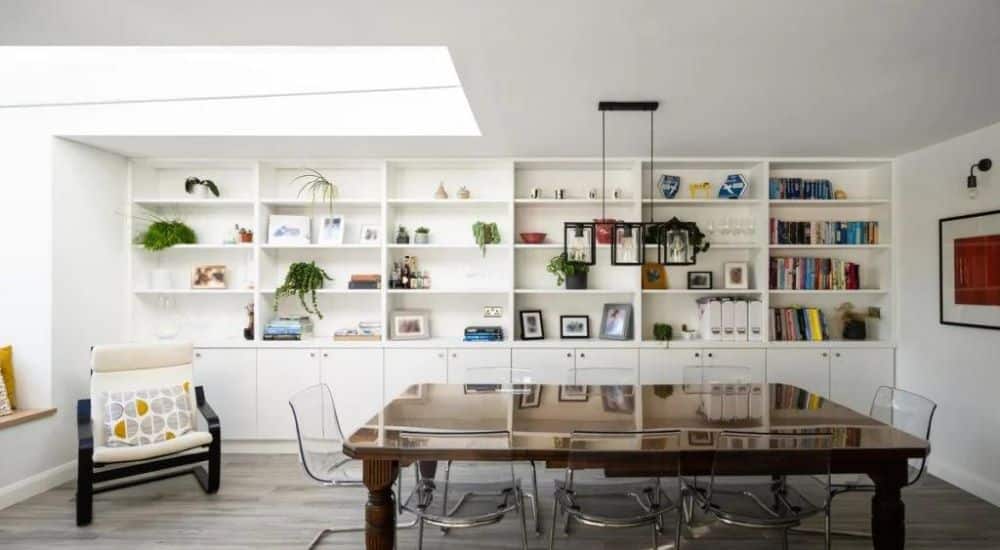 stretch over rooflight installed in a living and dining area, a large bookcase covers the furthest wall near a comfortable chair and a wooden dining table, surrounded by clear plastic chairs