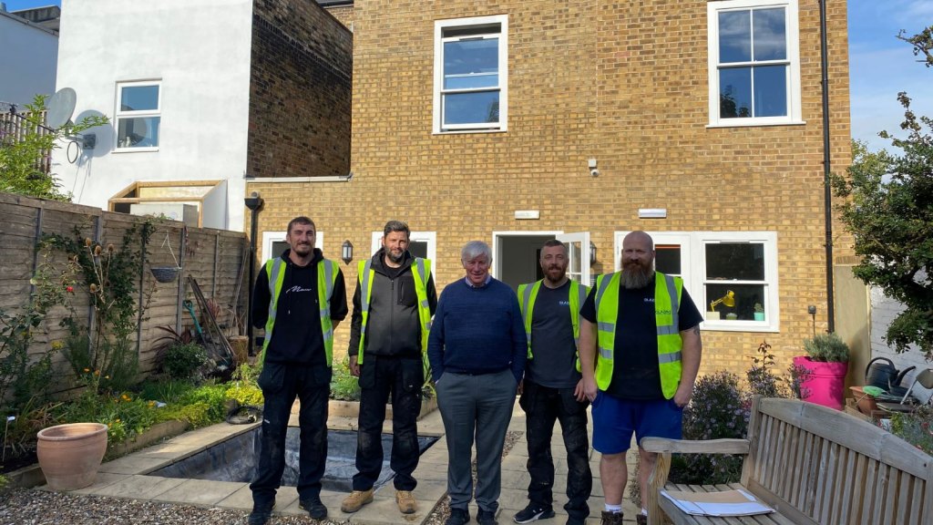 Two men in hi-viz jackets either side of a man in a blue jumper and grey trousers, they stand in front of a small office building