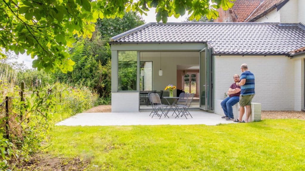 new extension of home with kitchen space can be seen through a bi folding door, two men sit on the garden wall surrounded by green grass and trees