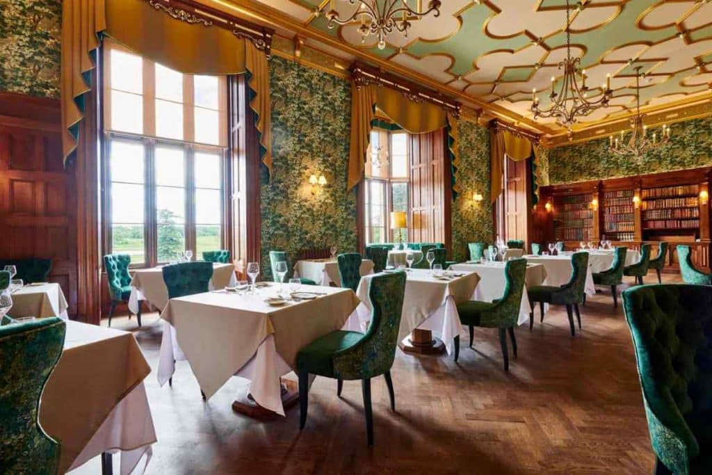 Dining room at hotel with green chairs at either end of small white tables, green patterned wallpaper surround windows