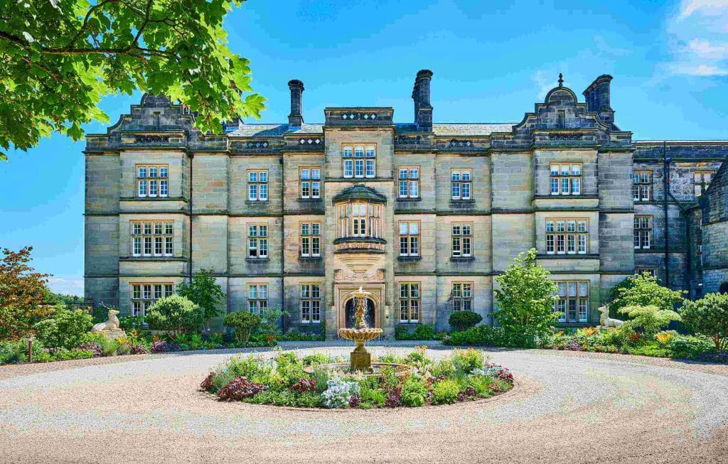 Exterior of historical hotel, Matfen Hall, surrounded by green bushes and trees, greenery and water feature in the center of the gravel pathway