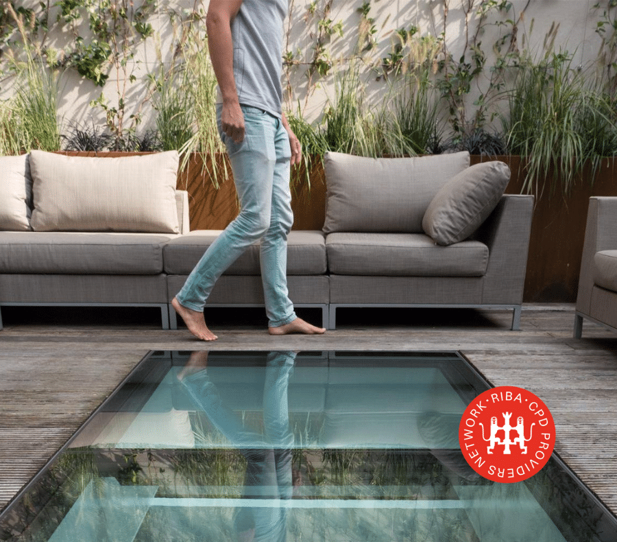 Man walking on garden patio decking past the outside sofa, walk on rooflight in foreground, RIBA Approved CPD logo in lower right corner